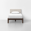 A single bed with a dark wooden frame, white bedding, and a beige upholstered headboard, positioned against a plain white wall in a minimal, modern room.