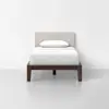 A single bed with a dark wooden frame, white bedding, and a beige upholstered headboard, positioned against a plain white wall in a minimal, modern room.