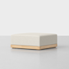 A modern, square ottoman with a light beige upholstered cushion and a wooden base, set against a plain white background.