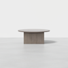 A minimalist round wooden table with a thick cylindrical base sits on a smooth, light-colored floor against a plain white background.