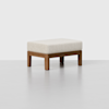 A rectangular wooden ottoman with light beige cushioned top, standing on four sturdy wooden legs, set against a plain light grey background.
