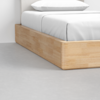 Close-up of a modern bed with a light wood frame and crisp white bedding, positioned on a smooth, light gray concrete floor.