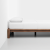A simple wooden bed frame with a white mattress, white sheets, and a white pillow, positioned against a plain white wall in a minimalistic room.