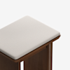 A close-up of a wooden stool with a light beige cushioned seat. The stool has a simple, modern design with clean lines and a smooth finish against a white background.