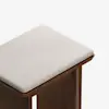 A close-up of a wooden stool with a light beige cushioned seat. The stool has a simple, modern design with clean lines and a smooth finish against a white background.