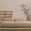 A minimalist living room with a beige, low-profile sofa, a wooden shelving unit holding a few books and decor items, and a tall potted plant against a plain light-colored wall.