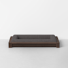 A modern, rectangular pet bed with a dark wood frame and dark gray cushioned sides, placed on a light gray floor against a plain white wall.