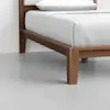 A close-up view of a wooden bed frame with clean lines, featuring white bedding and a pillow, set on a light grey floor.