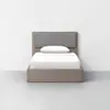A simple bed with a light wood frame, gray padded headboard, white bedding, and a single white pillow, placed against a plain white wall in a minimalist room.