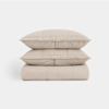 Two beige pillows are stacked on top of a folded beige comforter against a plain white background. All items have a quilted pattern and a soft, smooth texture.