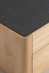 Close-up of a corner of a wooden cabinet with two drawers, featuring a smooth light wood finish and a dark, textured rectangular top surface.
