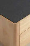 Close-up of a corner of a wooden cabinet with two drawers, featuring a smooth light wood finish and a dark, textured rectangular top surface.