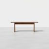 A simple wooden bench with a light-colored seat and darker legs, positioned against a plain white background on a smooth, light gray floor.