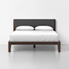 A modern bed with a dark wooden frame, gray upholstered headboard, white bedding, and two white pillows, positioned against a plain white wall on a light gray floor.