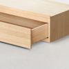 A close-up view of a light wood drawer partially open, showing smooth, minimalist construction and clean lines, set against a plain light gray floor.