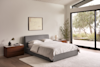A modern bedroom with a gray upholstered bed, white bedding, two wooden nightstands, a large window with a garden view, a plant, and a minimalist painting on the wall. Neutral tones create a calm, airy atmosphere.