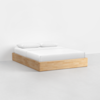 A minimalist bed with a light wood platform base, dressed in white sheets and two white pillows, set against a plain white wall and a light gray floor.