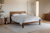 A minimalist bedroom with a wooden bed frame, white bedding, a light area rug, a bedside table with a lamp and decor, a tall potted plant, a large neutral artwork, and soft beige walls.