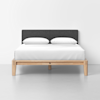 A modern bed with a light wood frame, dark gray upholstered headboard, and white bedding with two white pillows, set against a plain white wall and gray floor.