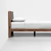 Side view of a modern wooden bed frame with a gray upholstered headboard, white pillow, and neatly made white bedding in a minimalistic, bright room.