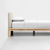 A minimalist wooden bed frame with a beige upholstered headboard, made up with white bedding and a single pillow, set against a plain white wall and floor.