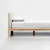 A minimalist wooden bed frame with a beige upholstered headboard, made up with white bedding and a single pillow, set against a plain white wall and floor.