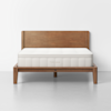 A wooden bed frame with a matching headboard supports a white, quilted mattress. The bed is centered against a plain white wall on a light gray floor. The word THUMA is visible on the front of the mattress.