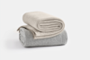 Two neatly folded, textured blankets are stacked on a white surface; the top blanket is light beige, and the bottom blanket is light gray with a subtle striped pattern.