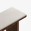 A close-up of a bench with a light beige upholstered cushion and a wooden frame, photographed against a plain white background.