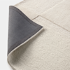 A close-up of a textured, light-colored area rug with a corner folded over, revealing a dark gray non-slip backing. The rug features subtle, geometric patchwork patterns in various shades of cream and beige.