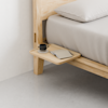 A minimalist wooden bedside table attached to a bed frame holds a black coffee cup, saucer, and two closed beige notebooks. The bed has light-colored bedding and the room features a clean, modern design.