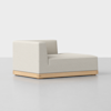 A modern, low-profile chaise lounge with light beige upholstery and a light wood base, featuring a single armrest on the left side, set against a plain white background.
