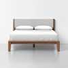 A modern wooden bed frame with a light gray upholstered headboard, white bedding, and two white pillows, set against a plain white wall and floor.