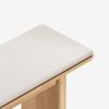 A close-up view of the corner of a wooden bench with a light-colored padded cushion on top, set against a white background.