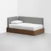 A modern daybed with a wooden base, white mattress, and gray upholstered backrest is placed in the corner of a minimalist room with white walls and a light gray floor.
