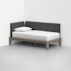 A minimalist daybed with a light wooden frame, white mattress, single white pillow, and dark gray upholstered backrest, placed in the corner of a white room with a gray floor.