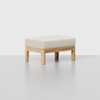 A minimalist ottoman with a light wood frame and a rectangular, cushioned top upholstered in a light beige fabric, set against a plain light background.