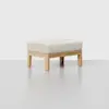 A minimalist ottoman with a light wood frame and a rectangular, cushioned top upholstered in a light beige fabric, set against a plain light background.