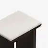 A close-up view of a wooden stool with a light beige, cushioned seat and dark brown legs, set against a white background.