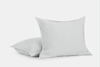 Two light gray pillows with a soft, smooth texture are placed on a white background; one pillow is standing upright while the other lies flat behind it.