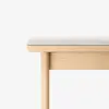 A close-up of a light wood bench with a rectangular beige cushion on top, shown against a white background. The bench has simple, clean lines and a minimalist design.