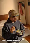 A person wearing glasses, a cap, and a sweatshirt sits on a bed holding a bowl of colorful cereal with a spoon. Clothes hang in the background. Text reads, Thuma + Heller Max Breakfast Set.