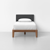 A single bed with a dark gray upholstered headboard, white bedding, and a single white pillow. The wooden bed frame has a minimalist design and sits on a light gray floor against a plain white wall.