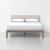 A modern bed with a light beige upholstered headboard, white bedding, and two white pillows. The bed frame is wooden with a natural finish, and the setting is a minimal, clean room with a light gray floor and white walls.