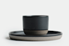A minimalist black ceramic cup sits on a matching round plate with a matte gray and black finish, set against a white background.