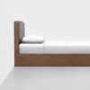 A minimalist wooden bed with a gray padded headboard, white pillow, and white bedding, positioned against a white wall and placed on a light gray floor.