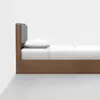 A minimalist wooden bed with a gray padded headboard, white pillow, and white bedding, positioned against a white wall and placed on a light gray floor.