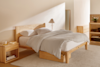A minimalist bedroom with a light wooden bed frame, white bedding, a matching nightstand, a lamp, a floor mirror, a dresser, shelves, and a beige rug on a wooden floor.
