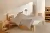 A minimalist bedroom with a light wooden bed frame, white bedding, a matching nightstand, a lamp, a floor mirror, a dresser, shelves, and a beige rug on a wooden floor.