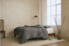 A minimalist bedroom with a bed featuring gray bedding, a wooden frame, a black floor lamp, a large window, a ceramic vase with branches, and neutral decor on a light carpeted floor.
